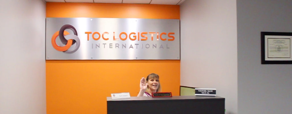 An individual sitting at a desk smiles and waves with a TOC Logistics International sign behind them.